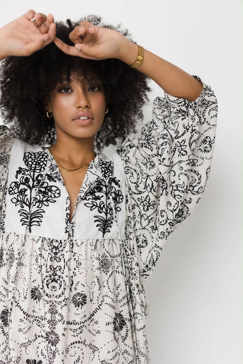 A woman with a large dark curly wig wearing a white long-sleeve bohemian tunic dress with black floral embroidery and paisley patterns, posing against a light gray background.