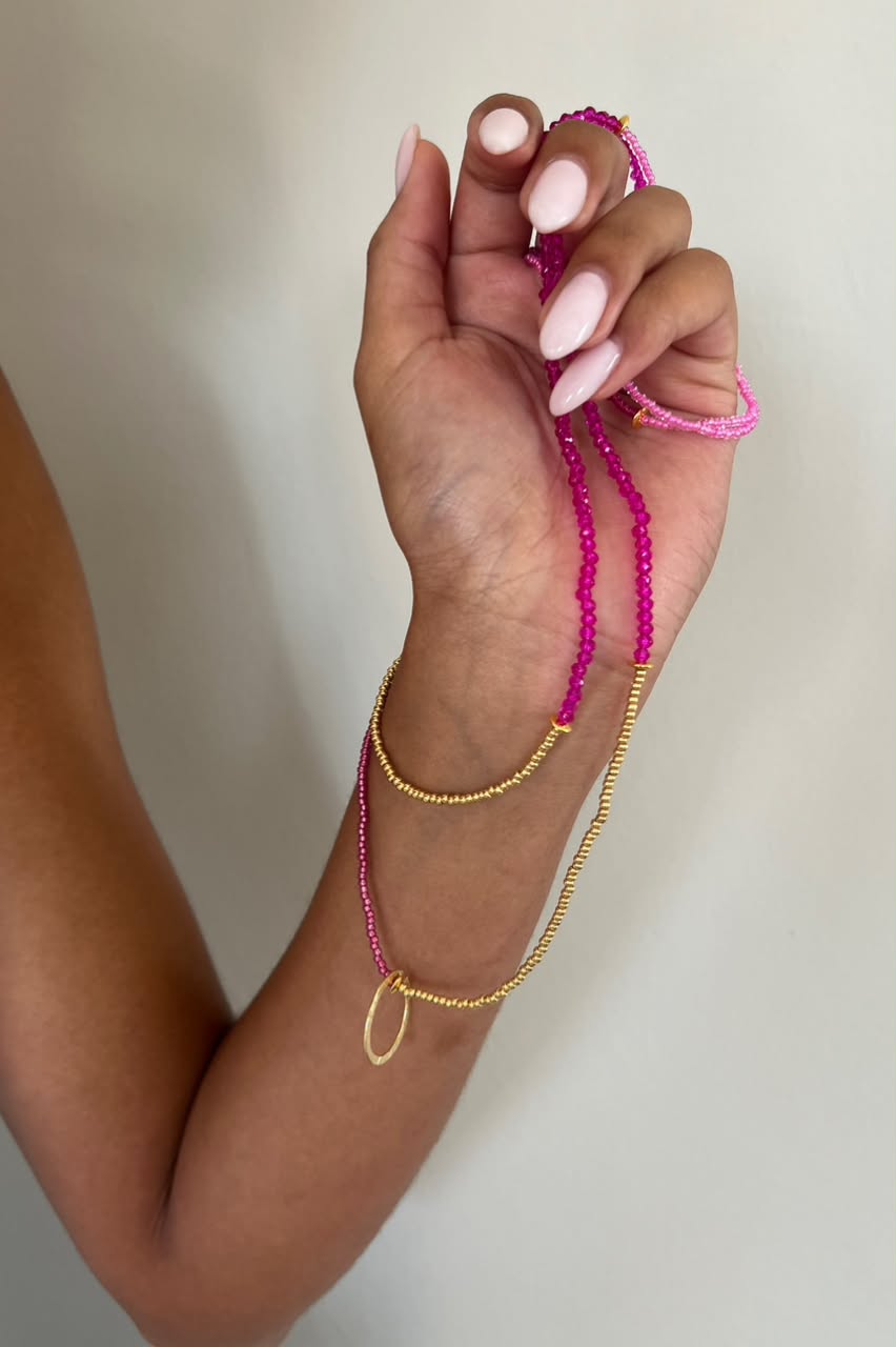 A close-up of a person's hand and wrist wearing a layered jewelry set featuring a bright pink beaded necklace with a gold ring pendant and a gold beaded chain.