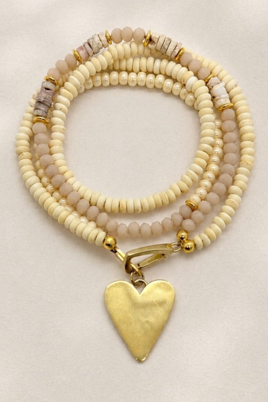 A multi-strand beaded bracelet with cream, beige, and light pink beads, accented with gold spacer beads and a textured gold heart-shaped pendant, displayed on a neutral cream background.