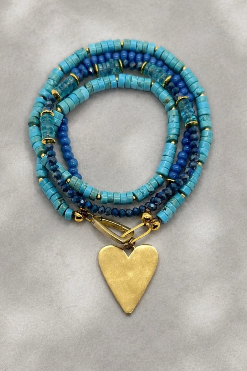 A multi-strand beaded bracelet featuring turquoise, royal blue, and navy beads with small gold spacer rings and a textured gold heart-shaped pendant, displayed on a neutral gray background.