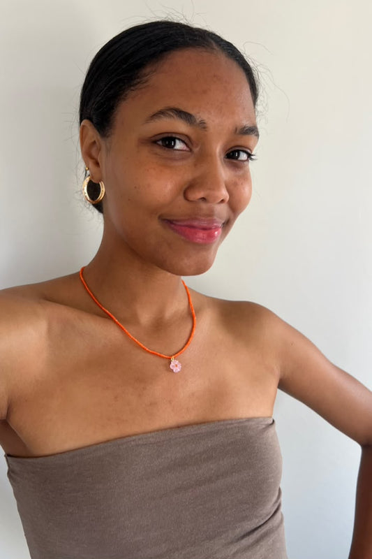 A woman wearing a thin orange beaded 
necklace with a small pink flower charm, paired with a taupe strapless top and gold hoop earrings, against a plain off-white wall.