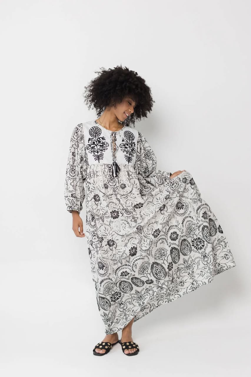 A woman with an afro hairstyle models a long-sleeved, loose-fitting maxi dress featuring a black and white botanical and paisley print. She is wearing black slide sandals and posing against a solid light gray studio background.