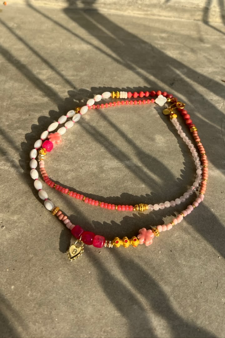 Two beaded necklaces with pink, coral, and white beads, accented with gold spacer beads and charms, displayed on a concrete surface with dappled sunlight and shadows.