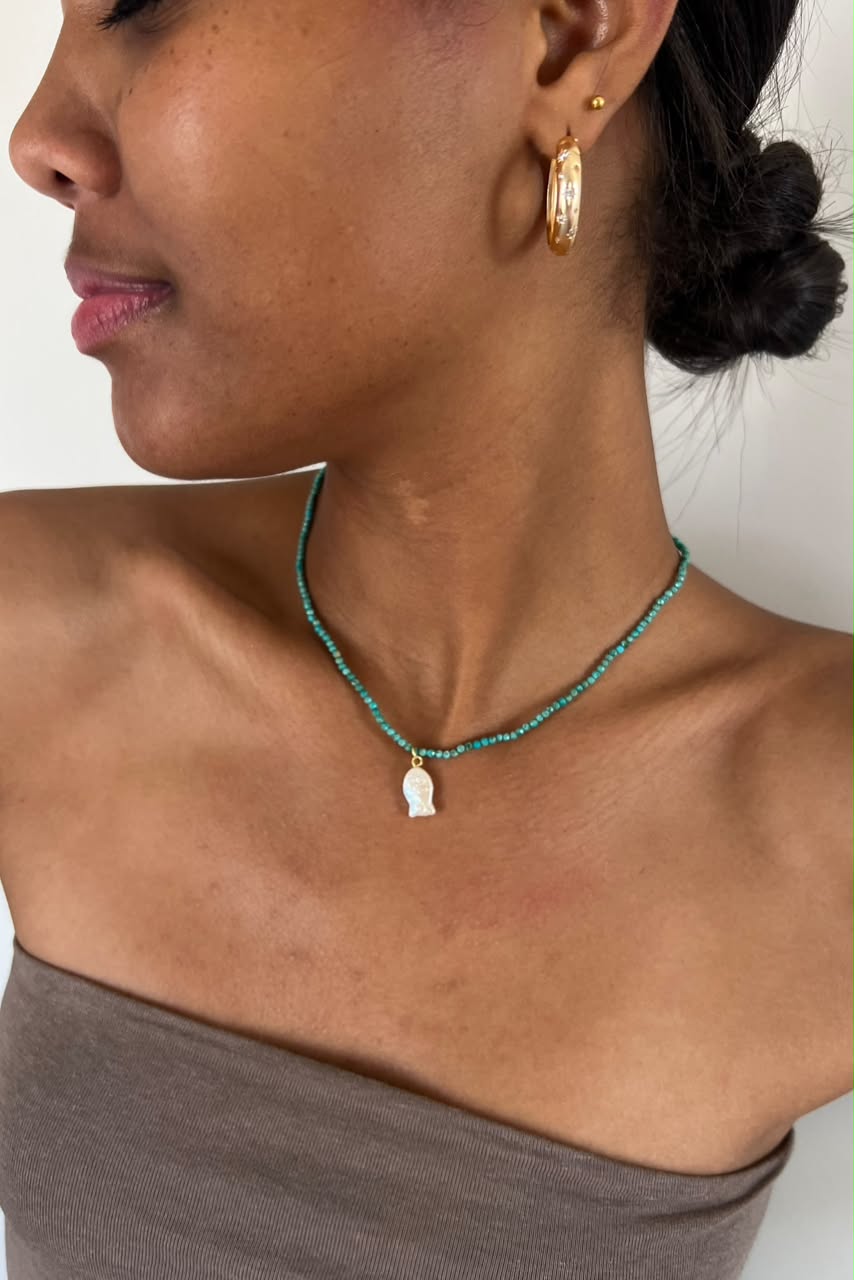 A close-up of a woman wearing a delicate beaded necklace with a small white pendant and gold hoop earrings, paired with a taupe strapless top.