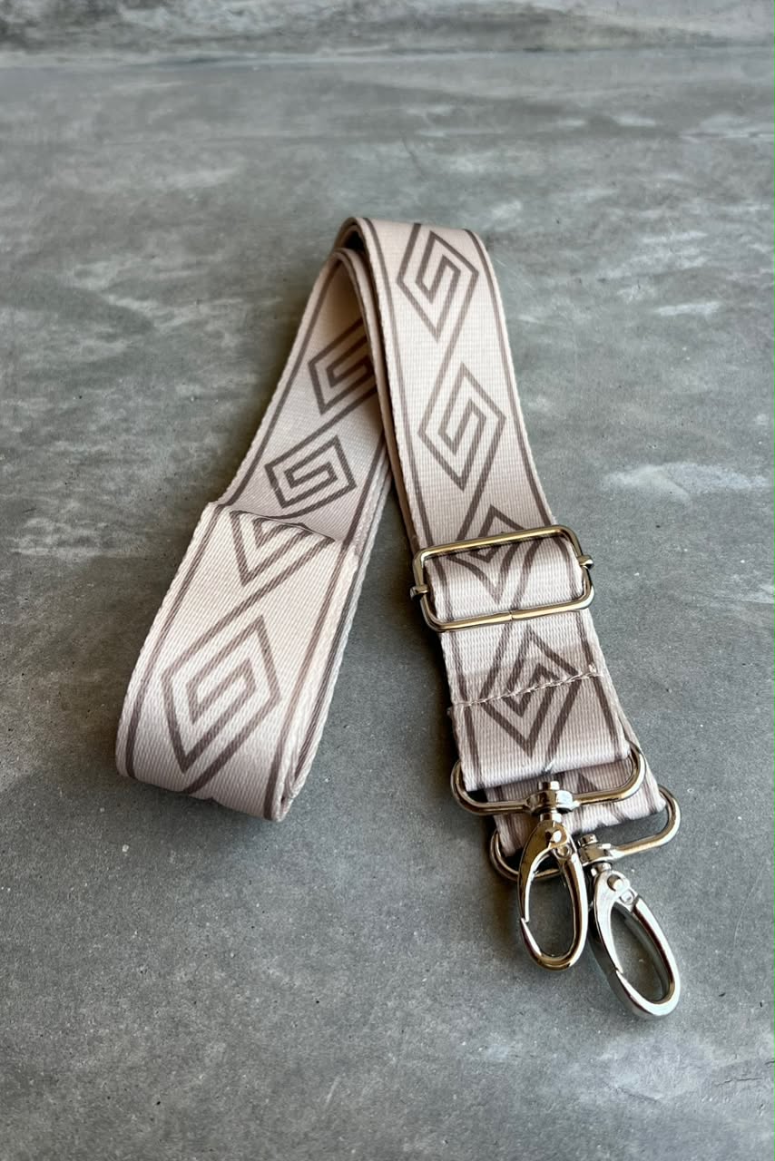A beige fabric shoulder strap with a dark brown geometric pattern, featuring a metal buckle and silver-toned swivel clips, displayed on a gray textured surface.