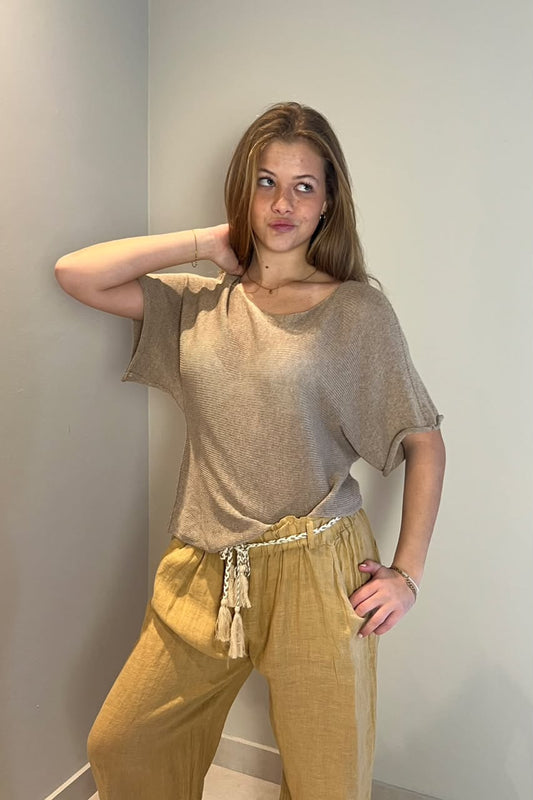 A woman wearing a beige short-sleeve knit top and linen pants, posing against a light studio background.