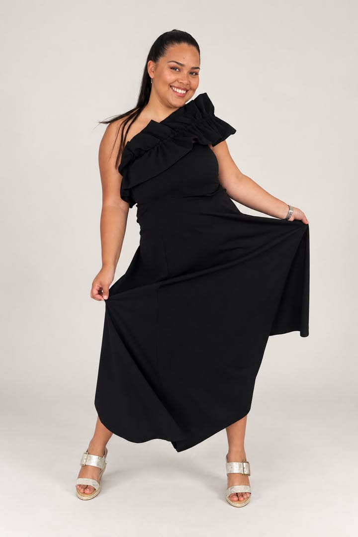 A woman modeling a black one-shoulder midi dress with a large ruffled shoulder detail and a flowing skirt, paired with metallic silver heeled sandals, against a light gray studio background.