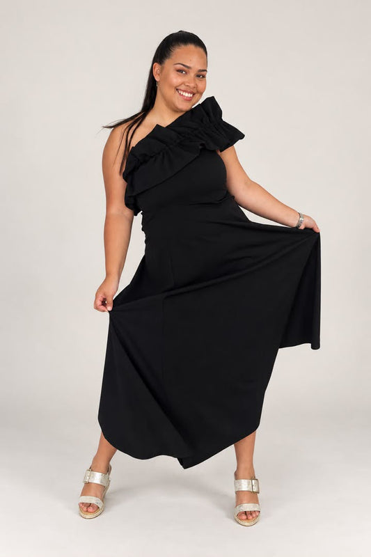 A woman modeling a black one-shoulder midi dress with a large ruffled shoulder detail and a flowing skirt, paired with metallic silver heeled sandals, against a light gray studio background.