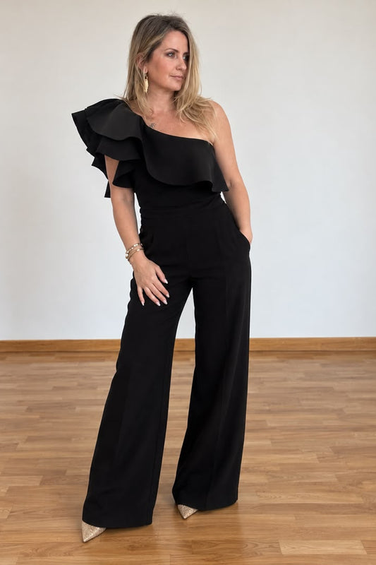 A woman modeling a black one-shoulder top with a dramatic ruffled sleeve and wide-leg pants, standing on a wooden floor against a plain light wall.