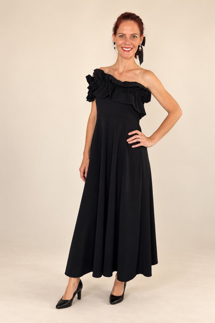 A woman modeling a black one-shoulder maxi dress with a ruffled shoulder detail and a flared skirt, paired with black heels, against a plain beige studio background.