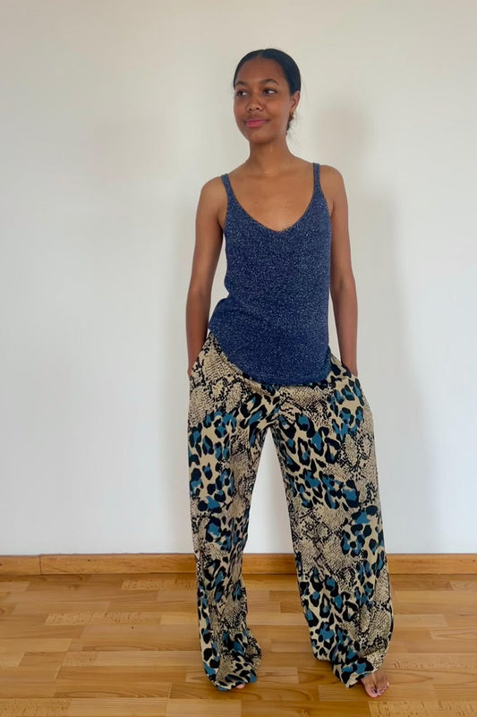 A woman stands barefoot on a light wood floor against a plain white wall, wearing a textured blue sleeveless top and loose patterned pants with a blue, black, and beige animal print.