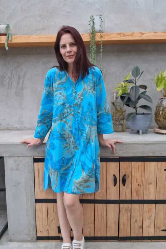 A woman with red hair wearing a bright blue button-up shirt dress with a green palm tree pattern, standing in front of a rustic concrete and wood counter with potted plants.