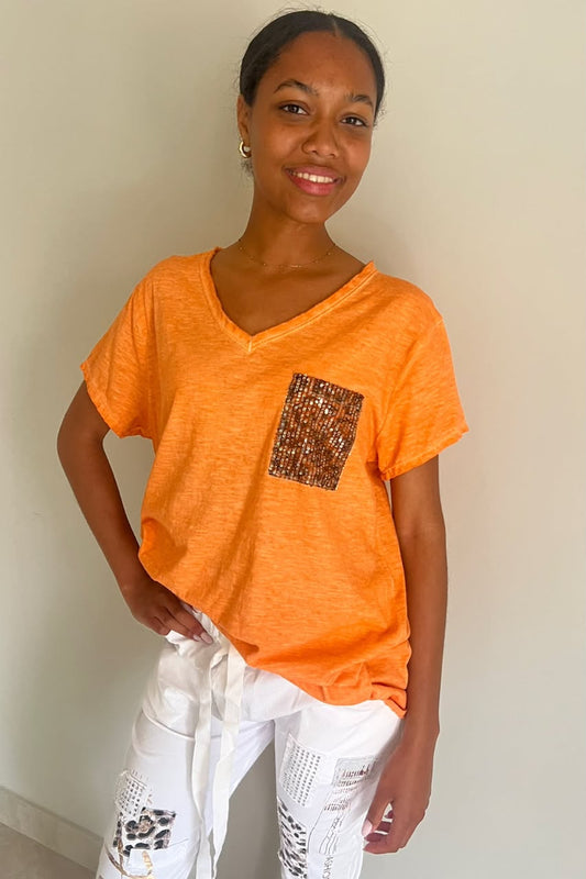 A woman wearing a bright orange V-neck t-shirt with a textured brown and gold patterned chest pocket, paired with white distressed pants, standing against a plain off-white wall.