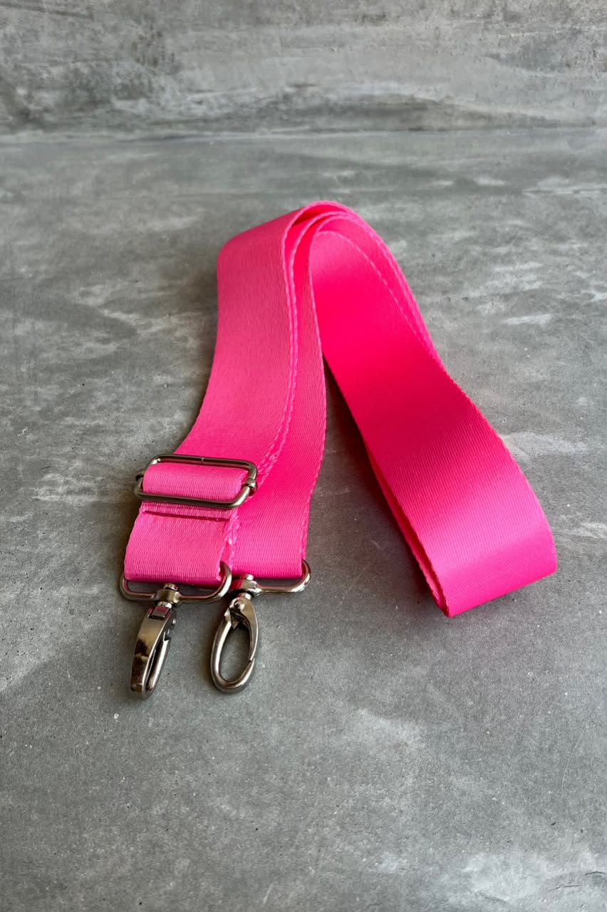 A bright pink fabric shoulder strap with silver metal hardware, including a quick-release buckle and two swivel clips, laid flat on a gray textured surface.