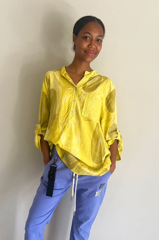 A woman wearing a bright yellow semi-sheer long-sleeve button-up shirt and light blue scrub pants, standing against a plain off-white wall.