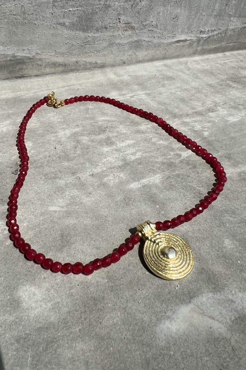 A beaded necklace featuring deep red faceted beads and a textured gold circular pendant, displayed on a gray textured surface.