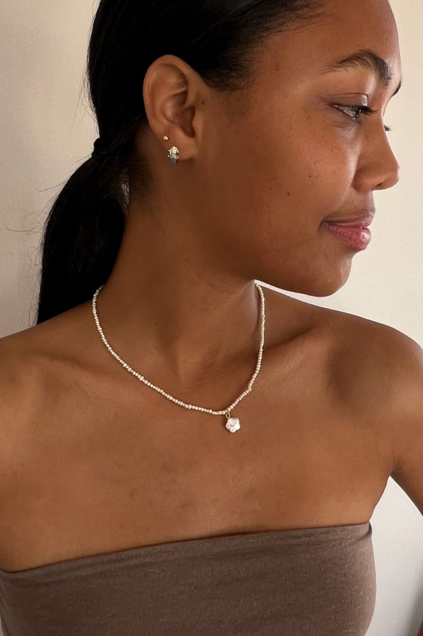 A woman in profile wearing a delicate white beaded necklace with a small pendant, paired with matching stud earrings, against a plain light background.