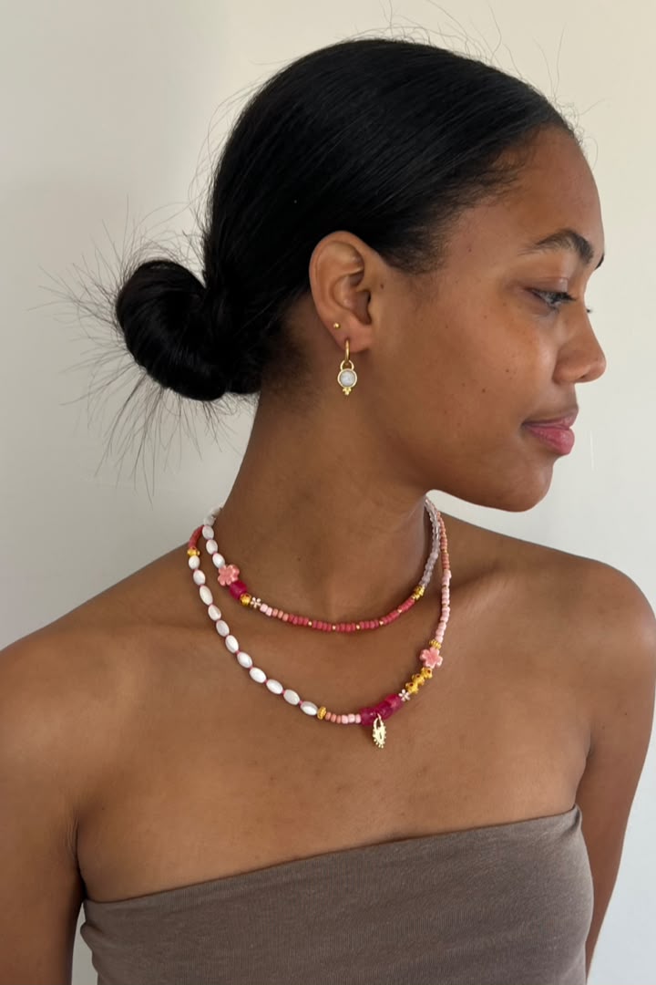 A woman in profile wearing a double-strand beaded necklace and matching drop earrings. The jewelry features pink, red, white, and yellow beads with gold-toned metal accents, including a small gold leaf charm on the necklace.