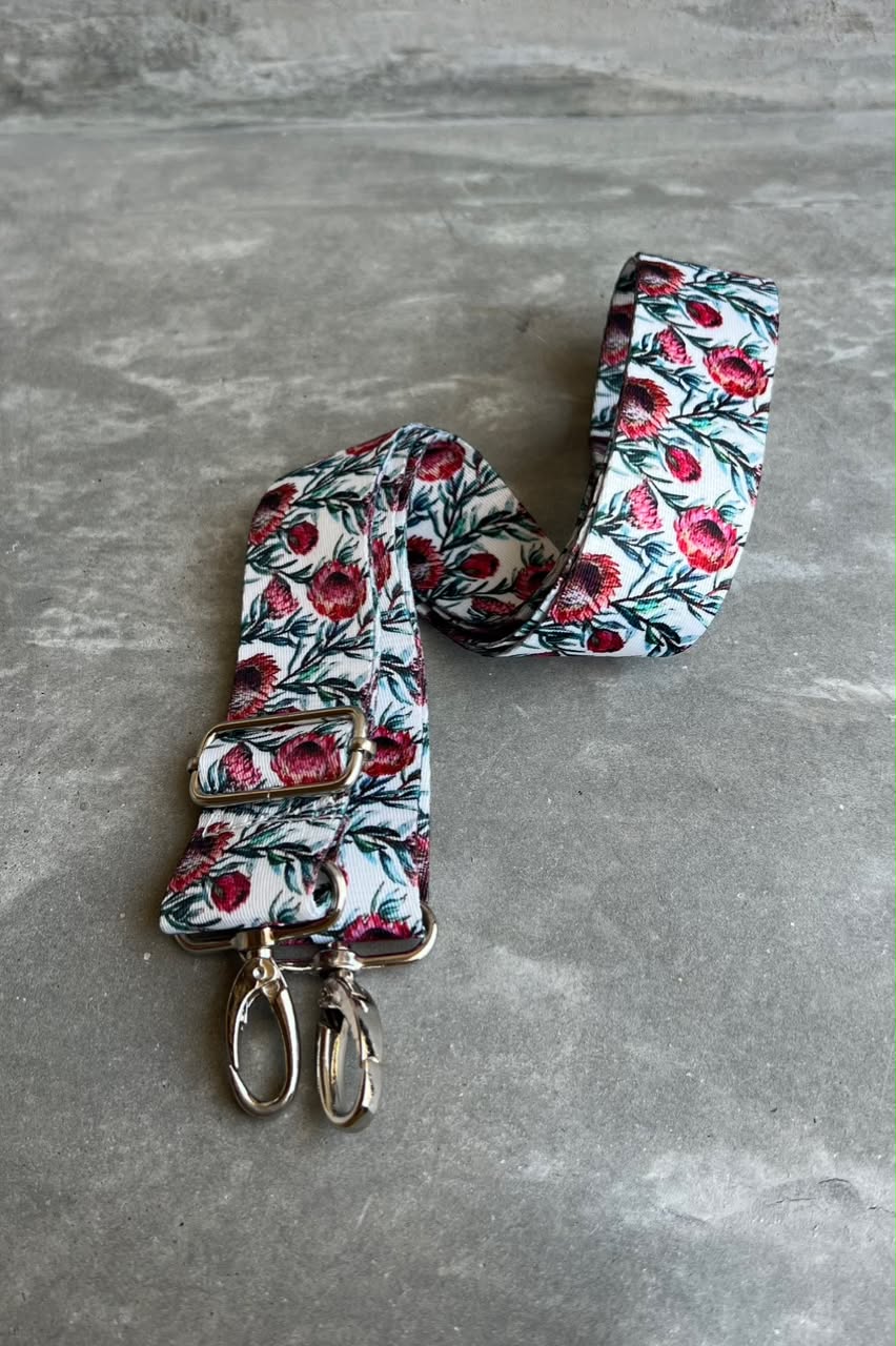 A floral fabric strap with a white background, red and pink flowers, and green leaves, featuring gold-tone metal clips at one end, displayed on a gray textured surface.