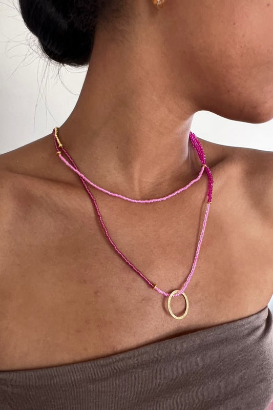 A close-up of a model's neck and upper chest wearing a layered necklace. The necklace features a top layer of light pink seed beads and a bottom layer of darker magenta-pink seed beads, both accented with small gold spacers and a gold circular pendant.