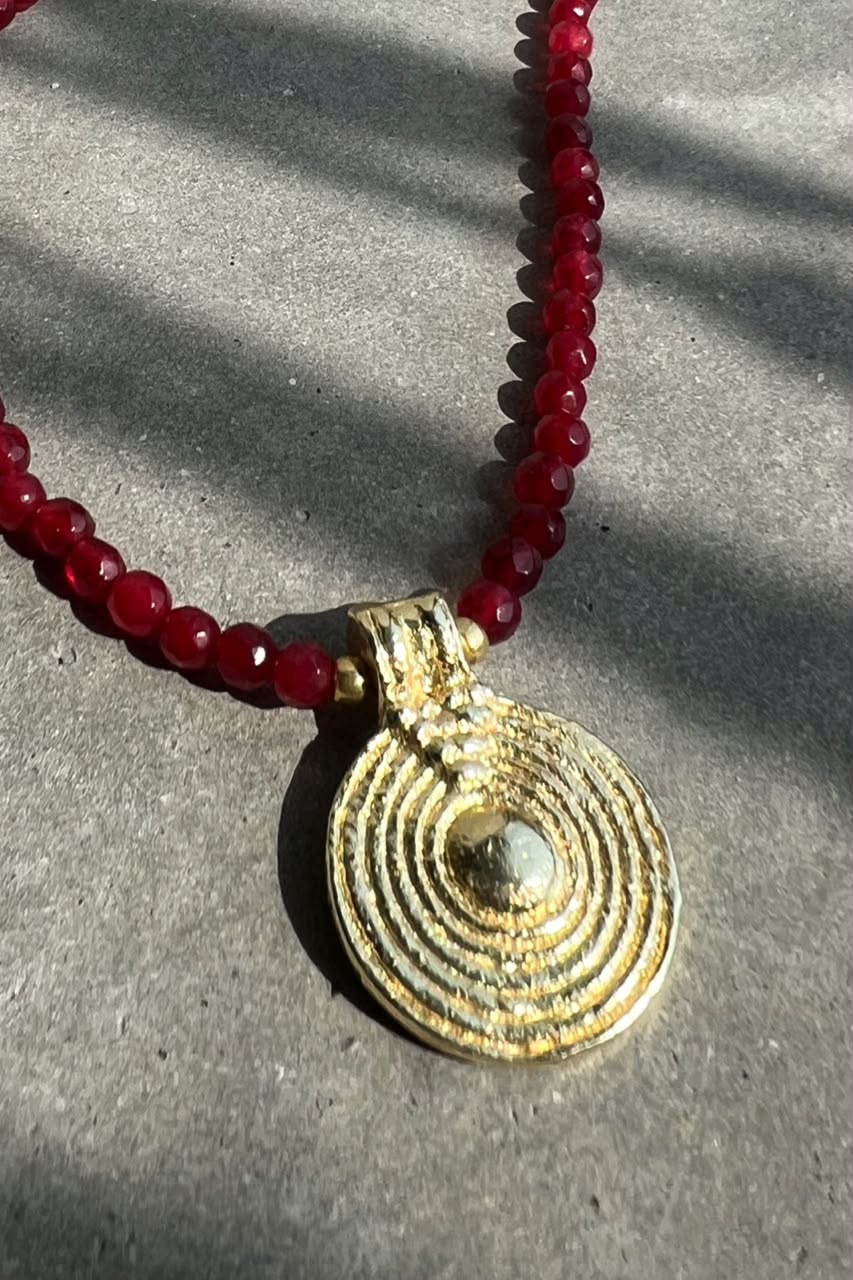 A close-up of a necklace with a textured gold circular pendant and a red beaded chain, resting on a gray surface with natural shadows.