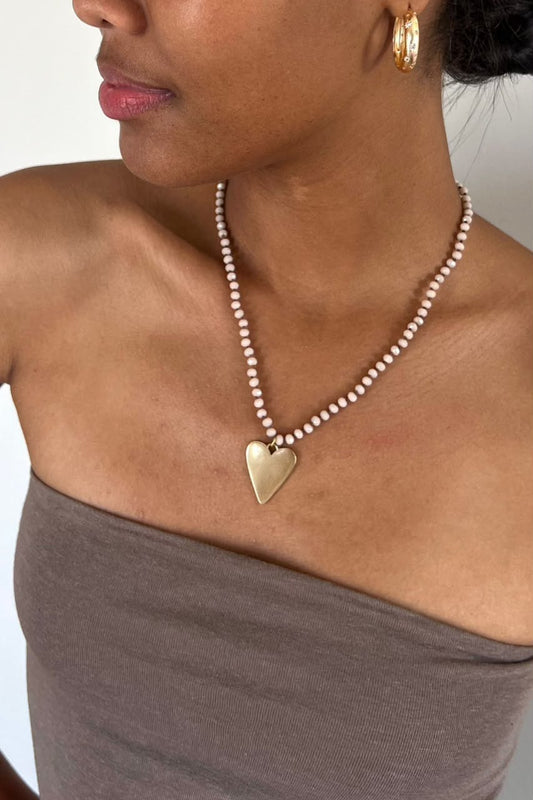 A close-up of a model wearing a delicate beaded necklace with a gold heart-shaped pendant, paired with a taupe strapless top.