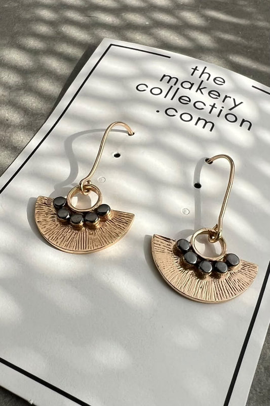 A pair of gold-toned dangle earrings with a fan-shaped textured design, small black bead accents, and curved hooks, displayed on a white card labeled 'the makery collection .com' under dappled sunlight.