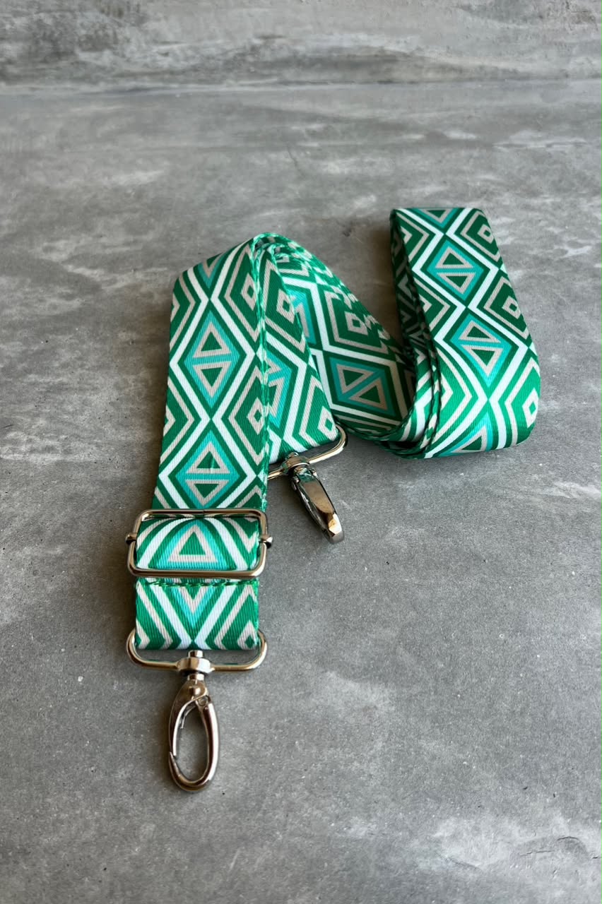 A green and white geometric patterned fabric strap with silver-tone metal hardware, including a buckle, clips, and a swivel ring, displayed on a gray textured surface.