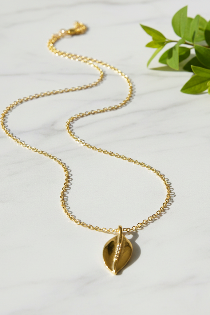 Gold necklace with sparkling leaf charm draped on marble surface with green leaves nearby