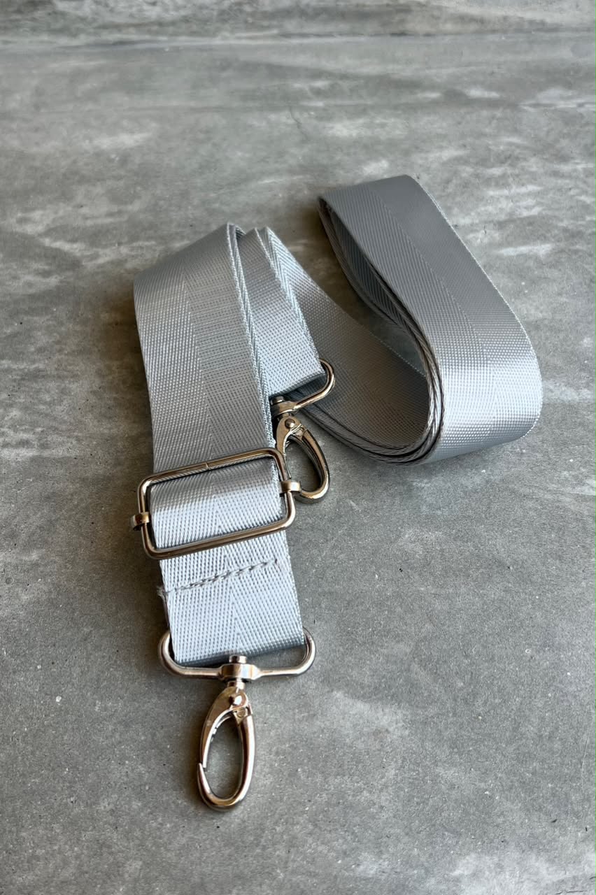 A light gray fabric strap with silver metal hardware, including a buckle, rings, and a swivel clasp, laid out on a gray textured surface.
