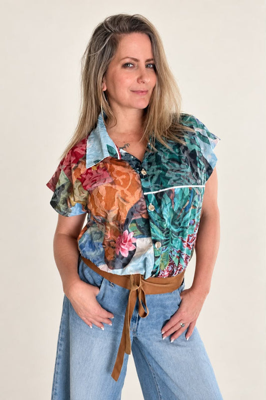 A woman modeling a short-sleeve button-up blouse with a multicolor floral and paisley print, paired with a brown belt and light blue jeans, against a plain beige background.