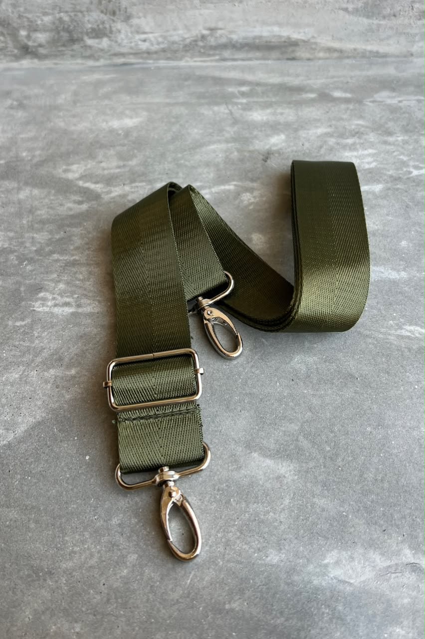 An olive green webbing strap with silver-tone metal hardware, including a buckle, swivel clips, and a loop, laid out on a textured gray surface.