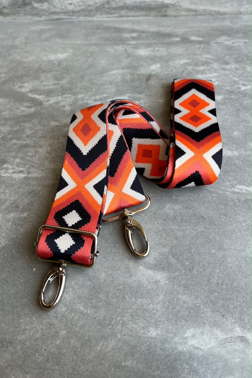 A woven fabric shoulder strap with a geometric Southwestern pattern in orange, black, white, and red, featuring silver-tone metal swivel clips on both ends, displayed on a gray textured surface.