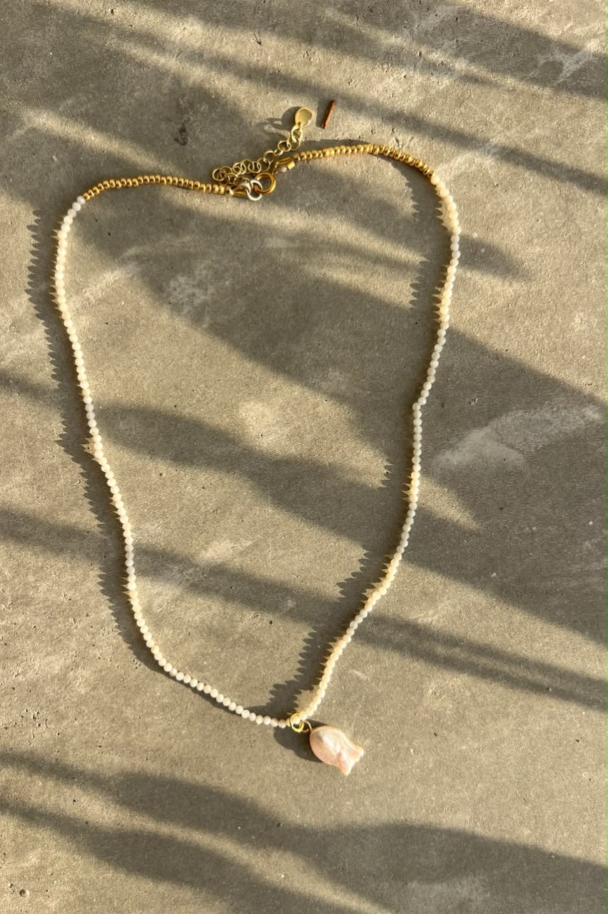 A delicate pearl chain necklace with a small light pink shell pendant, laid flat on a textured gray concrete surface with dappled sunlight and shadows.