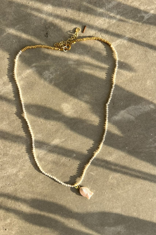 A delicate pearl chain necklace with a small light pink shell pendant, laid flat on a textured gray concrete surface with dappled sunlight and shadows.