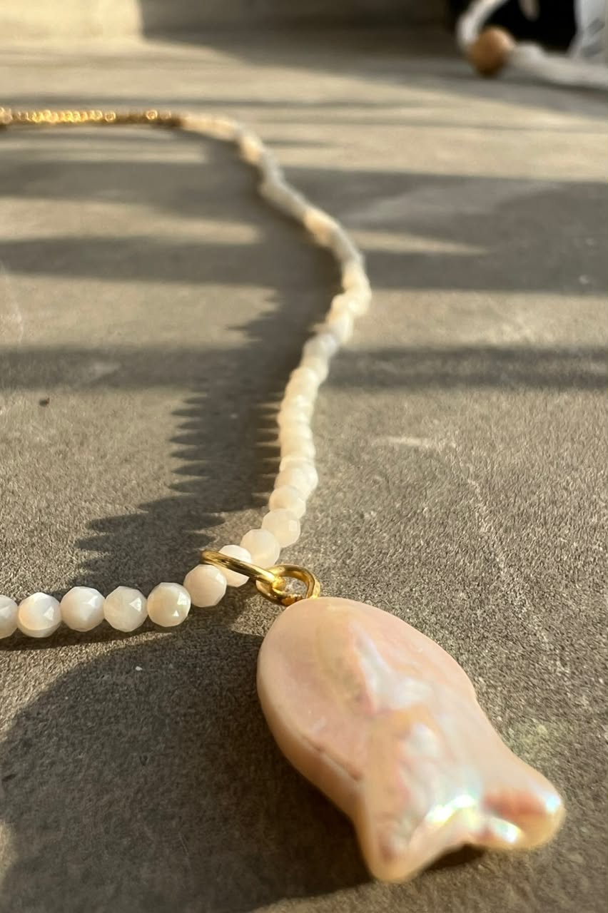 A close-up of a necklace laid on a textured gray surface in bright sunlight. The necklace features a pearl pendant with a gold clasp, attached to a strand of small white pearls.