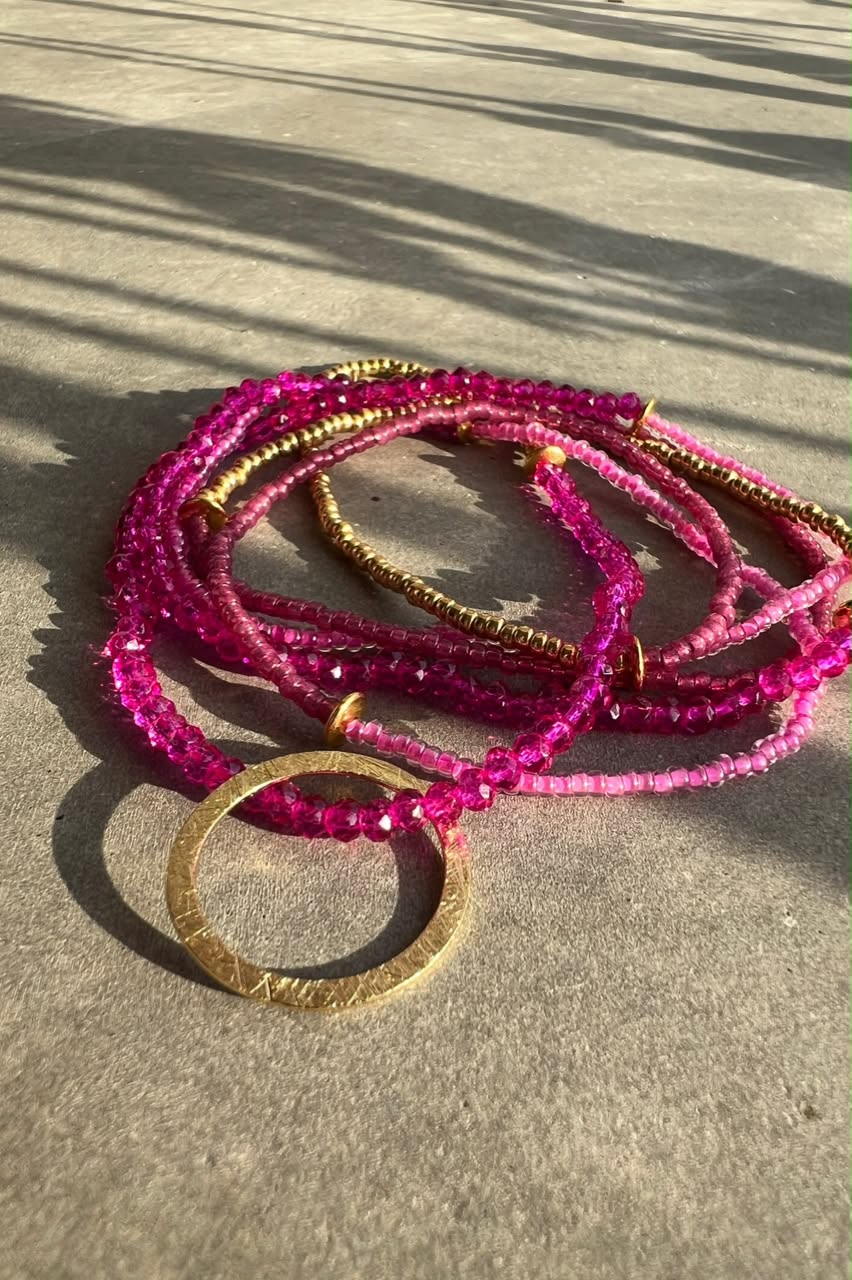 Multi-strand beaded bracelets in vibrant pink and metallic gold, featuring a large textured gold ring, arranged on a concrete surface under direct sunlight.