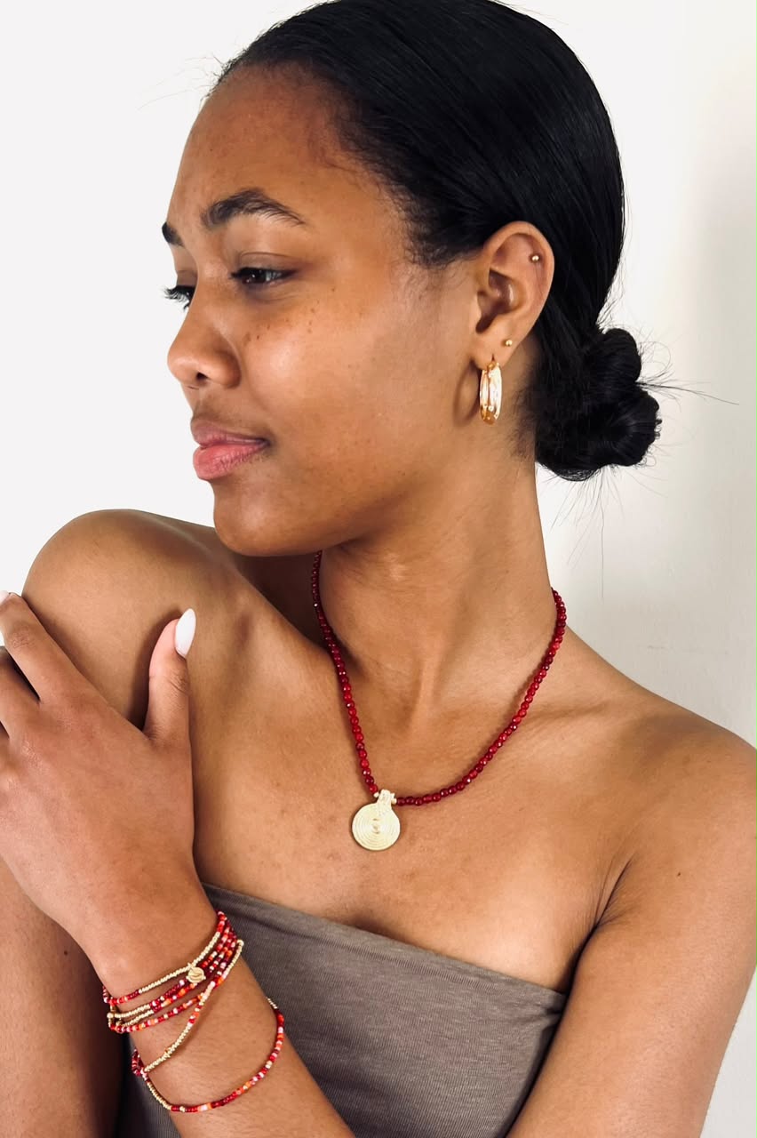 A woman wearing a red beaded necklace with a round gold pendant, matching red and gold bracelets, and a gold hoop earring, shown in a close-up portrait against a plain light background.