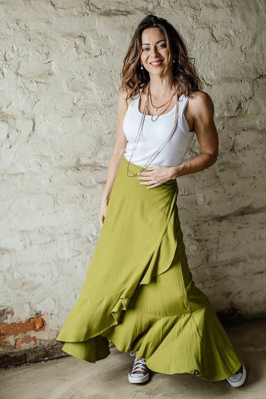 A woman wearing a white tank top and a flowing green ruffled maxi skirt, standing against a rustic textured brick wall.