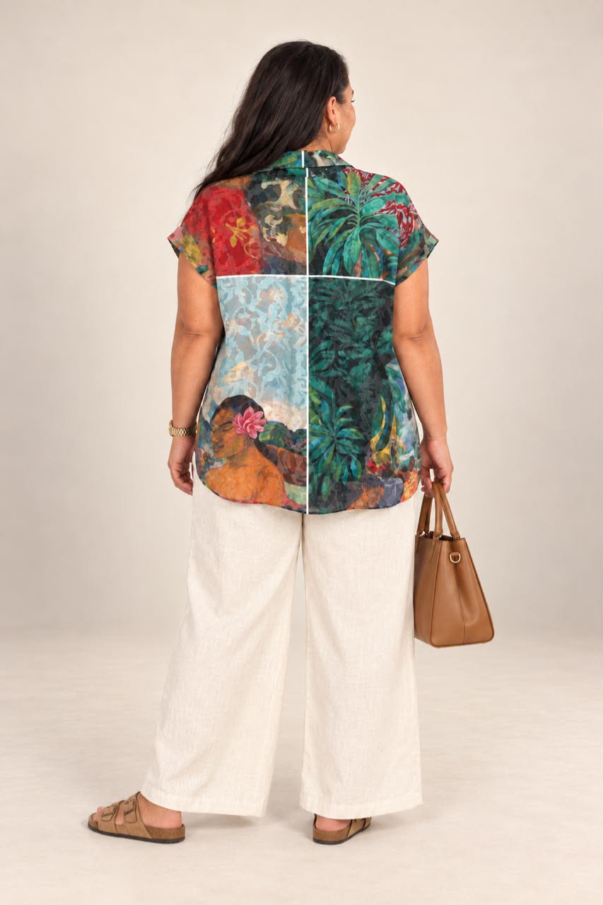A woman shown from the back wearing a short-sleeve button-up shirt with a colorful patchwork print of tropical foliage and floral scenes, paired with wide-leg white pants and brown sandals, holding a brown handbag against a plain beige studio background.