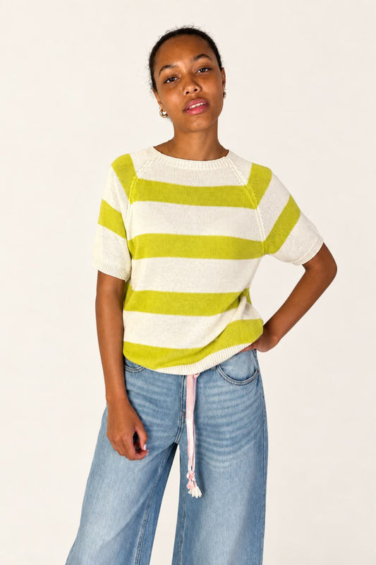 A woman wearing a short-sleeve knit top with horizontal lime green and off-white stripes, paired with light blue jeans, against a plain off-white studio background.