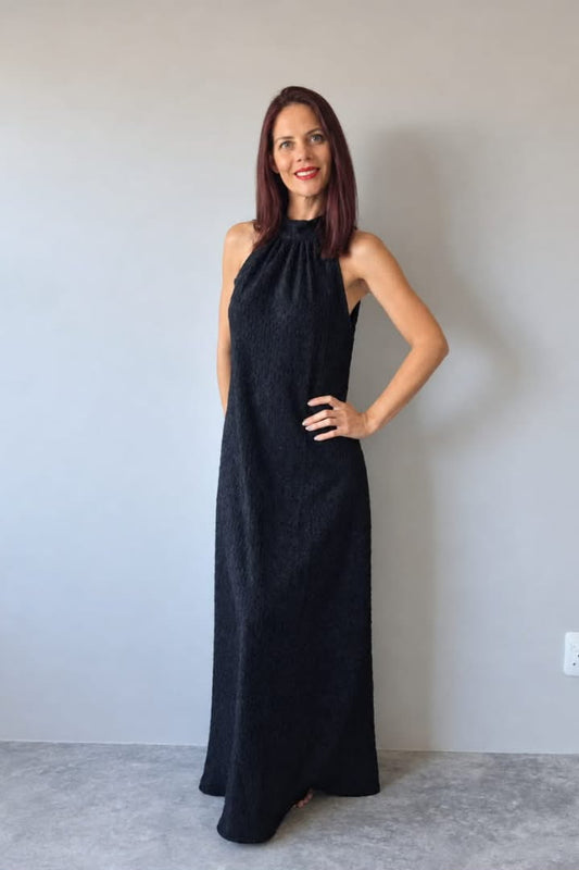 A woman modeling a sleeveless black halter-neck maxi dress with a textured pattern, standing against a plain light gray wall.