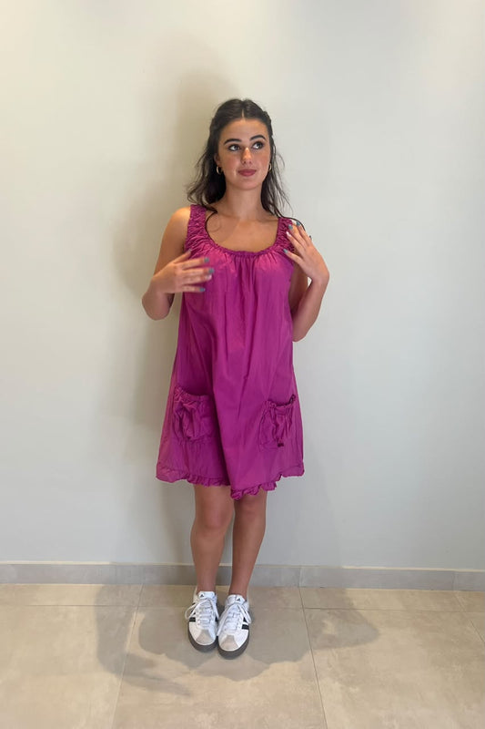 A woman modeling a bright magenta sleeveless dress with ruffled trim and side pockets, paired with white sneakers, standing against a plain light gray wall.