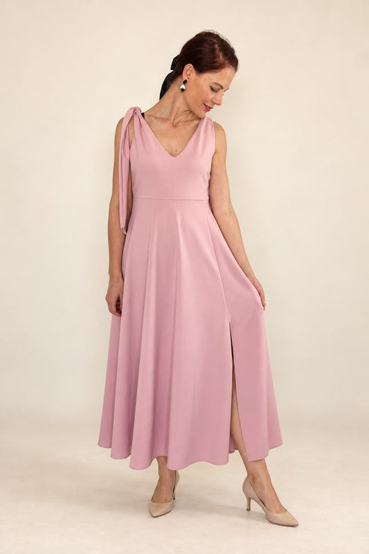 A woman modeling a sleeveless dusty pink maxi dress with a draped shoulder detail, V-neckline, and front slit, paired with beige heels against a plain cream studio background.