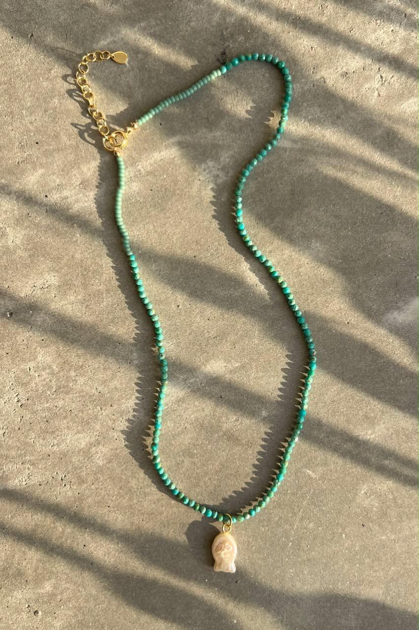 A beaded necklace with turquoise green beads, a gold-tone chain extender, and a small cream-colored pendant, laid out on a concrete surface with natural shadow patterns.