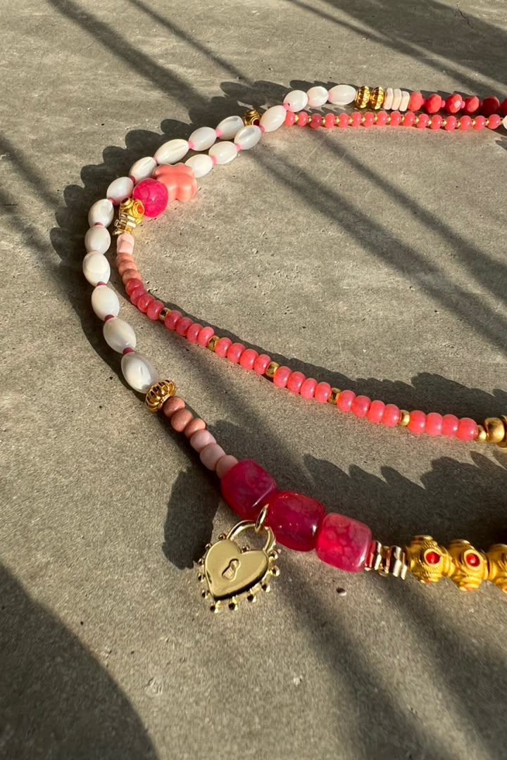 A beaded necklace with white and pink beads, gold spacer details, and a gold heart-shaped pendant, laid on a gray concrete surface with strong sunlight and shadows.