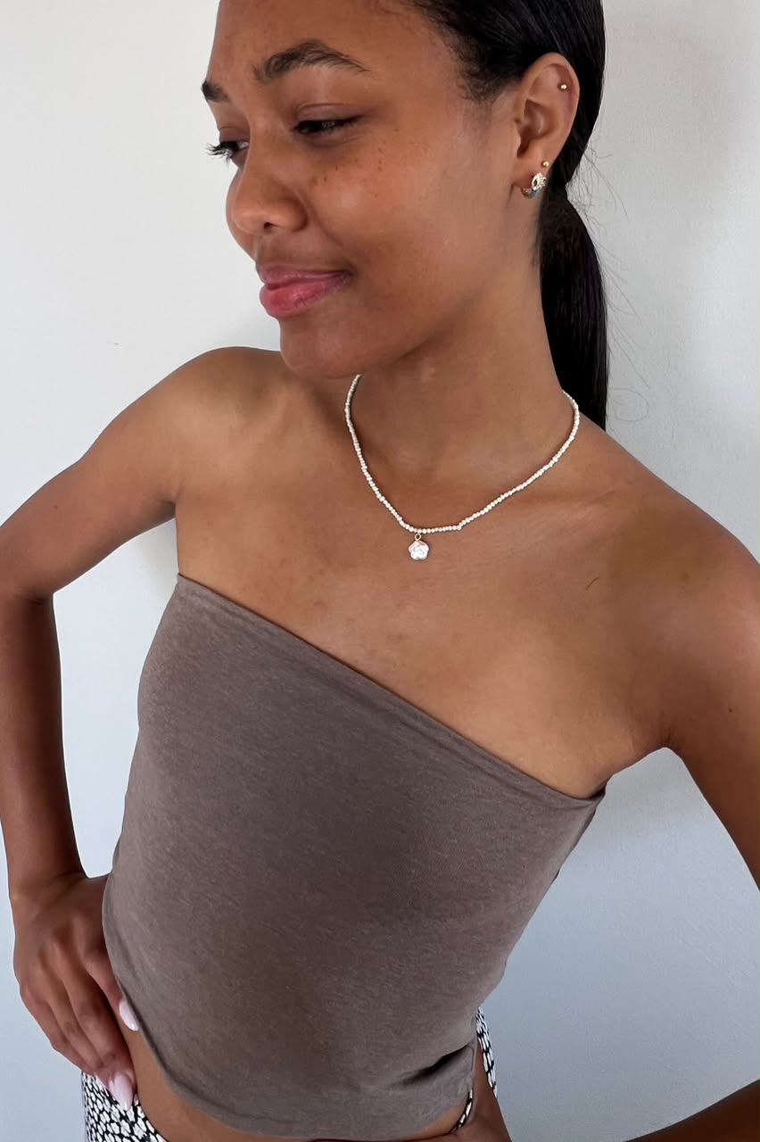 A woman wearing a taupe-brown strapless tube top and a delicate white necklace with a small heart pendant, shown from the waist up against a plain light gray wall.