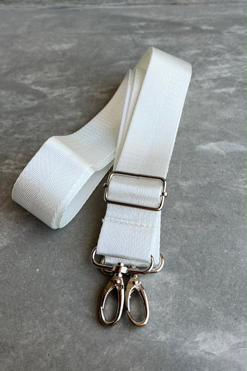 A white nylon webbing strap with silver metal hardware, including a buckle, loops, and two hanging clips, displayed on a textured gray surface.