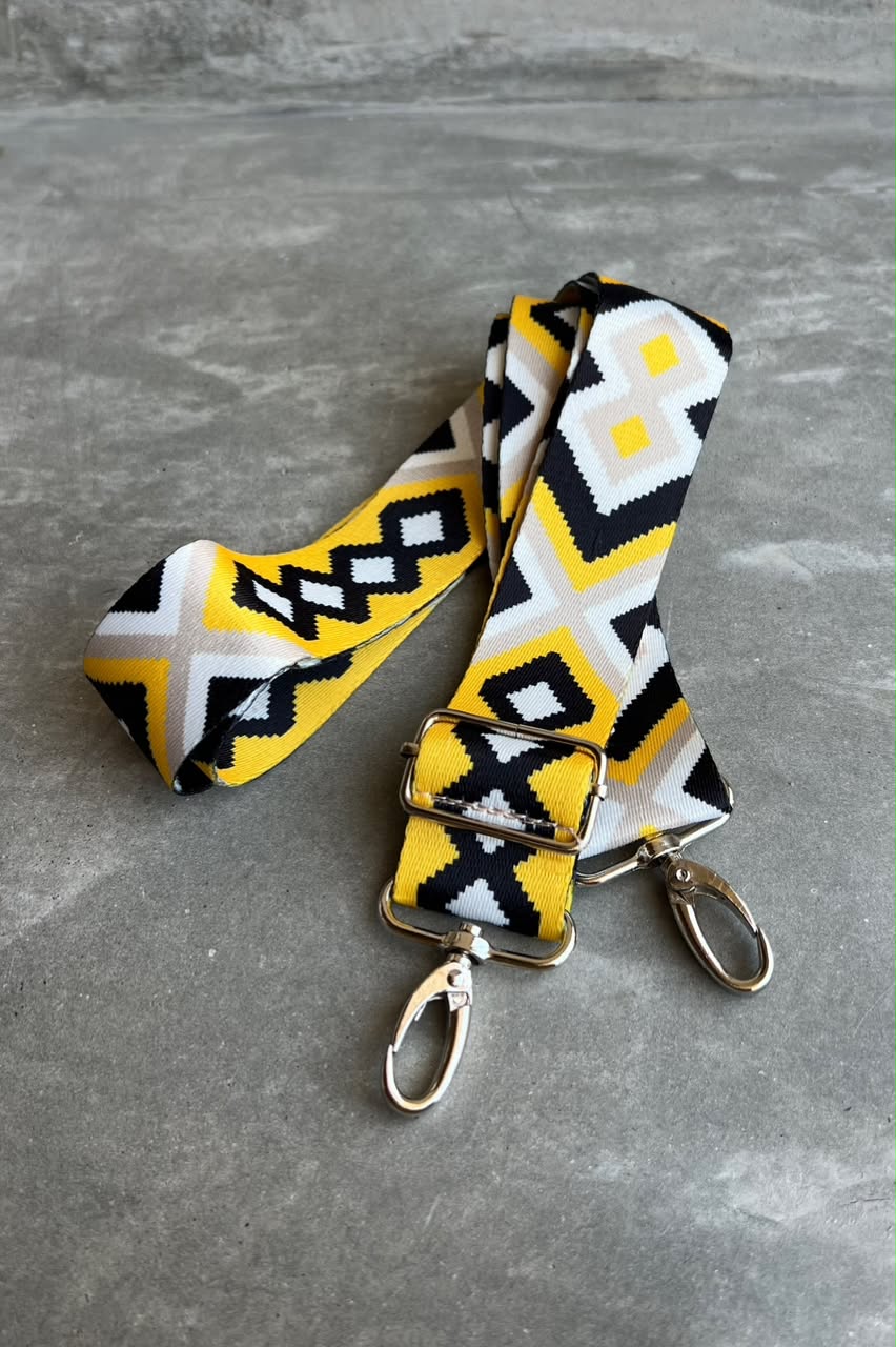 A woven fabric strap with a geometric pattern in yellow, black, and white, featuring silver metal hardware including a buckle, swivel clips, and a lobster clasp, displayed on a gray textured surface.